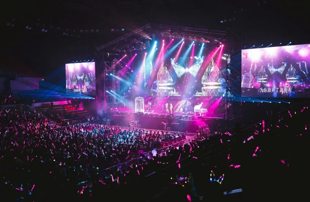 A packed arena concert with bright stage lights and giant screens above the crowd