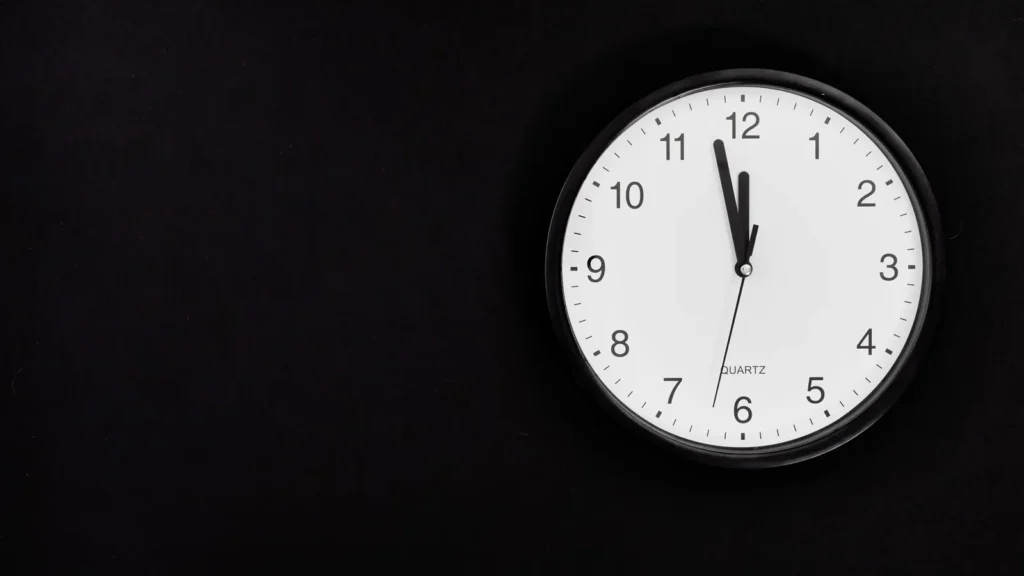 A plain wall clock against a dark background with its hands just before midnight