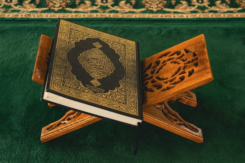 A copy of the Qur'an resting on an ornate carved wooden rehal on a green prayer carpet